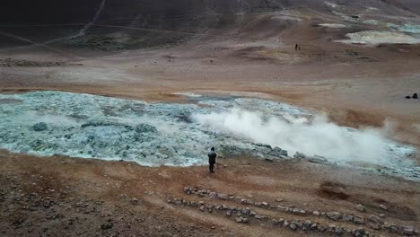 Aerial,-orbit,-drone-shot,-around-a-woman-taking-pictures-of-a-steaming-crater-emitting-sulfuric-gas,-at-Hverir-geothermal-area,-on-a-sunny,-summer-day,-in-North-Iceland