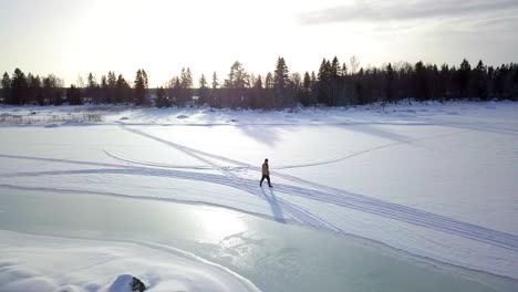 Aerial-descending,-tracking-shot-of-lonely-older-man-walking-on-icy-frozen-lake-in-winter,-in-Lapland,-Finland
