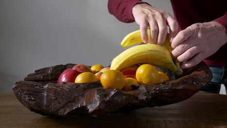 A-man-takes-a-banana-from-a-wooden-bowl-with-fruits-standing-on-a-wooden-table