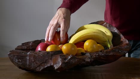 A-male-hand-takes-an-apple-from-a-wooden-bowl-with-fruits-standing-on-a-wooden-table