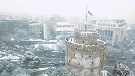 Aerial-footage-of-white-tower-of-Thessaloniki-during-snowstorm,-Greek-flag-waving-on-top-of-the-castle