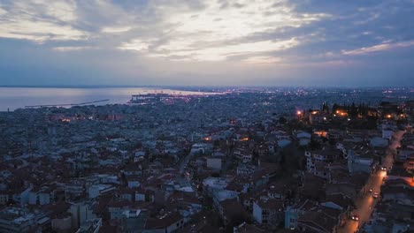 Aerial-drone-flying-over-romantic-old-town-city-of-Thessaloniki-by-night-illuminated,-Greece,-port-by-dusk