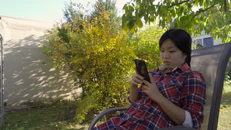 girl-in-garden-sits-comfortably-in-chair-reading-mobile,-low-angle-medium-shot