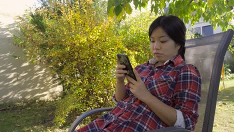 asian-lady-relaxes-in-chair-reading-messages-on-phone,-low-angle-medium-shot