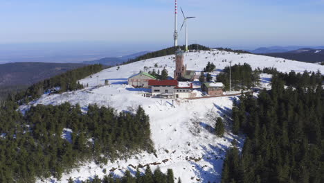 Aerial-shot-ascending-over-snow-covered-hill-showing-broadcasting-tower,-wind-turbine-and-buildings
