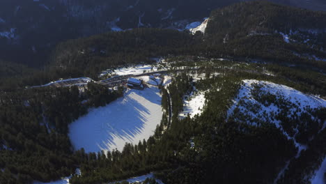 Aerial-shot-closing-in-to-a-frozen-black-forest-lake-and-hotel,-are-only-partly-snow-covered,-golden-hour-morning-light