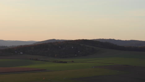 A-time-lapse-view-of-a-natural-airport-field-and-a-hill-where-one-can-see-several-smaller-houses-built-under-a-ridge-captured-during-the-sunny-autumn-sunset-area-of-Brno-Czech-Republic