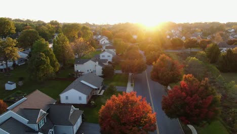 Sunset-starburst-highlights-autumn-neighborhood-scene-with-homes-and-colorful-trees,-cars-and-trucks-on-street-at-intersection,-aerial-drone-shot