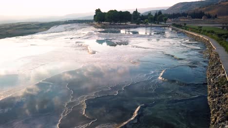 Drone-flight-over-Pamukkale-with-beautiful-reflections-of-sunset-and-clouds-in-the-water-and-white-salt-formations,-travertines-looking-over-valley-in-Turkey