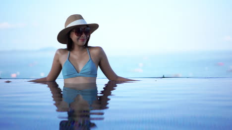 Fröhliche,-Stilvolle-Asiatische-Frau,-Die-Im-Badeanzug-Am-Rand-Des-Infinity-Pools-Mit-Sonnenbrille-Und-Sommerhut-Steht