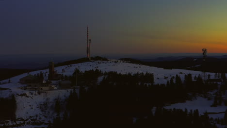 Aerial-shot-at-dawn-ascending-over-snow-covered-hill-showing-broadcasting-tower,-wind-turbine-and-buildings
