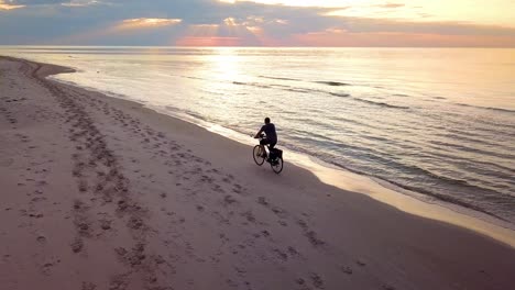 Aerial-Slowmotion-Shot-of-a-Young-Man-Riding-Bicykle-on-Beach-at-Sunset