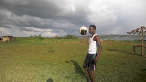 Slow-motion-wide-shot-of-a-young-African-man-doing-kick-ups-on-a-beach-by-a-lake-with-storm-clouds-rolling-in