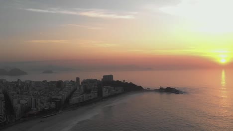 Luftschwenk,-Der-Den-Sonnenaufgang-über-Dem-Meereshorizont-Hinter-Arpoador-Zeigt,-Der-Den-Breiteren,-Leeren-Strand-Von-Ipanema-Am-Frühen-Morgen-Im-Vordergrund-Und-Das-Breitere-Stadtbild-Von-Rio-De-Janeiro-Im-Hintergrund-Zeigt