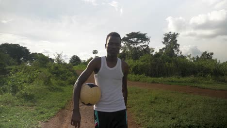 Front-view-tracking-a-young-African-man-walking-away-from-the-sun-with-a-football-under-his-arm