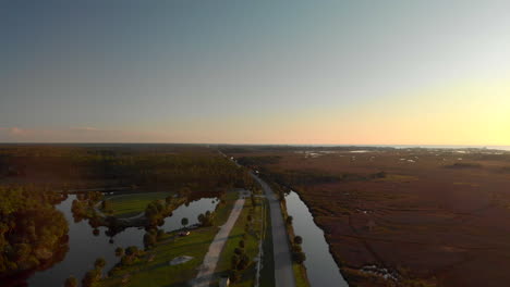Aerial-view-of-a-long-road-built-in-between-two-lakes,-with-some-cars-driving-towards-a-large-forest,-sunset-shot-in-Florida