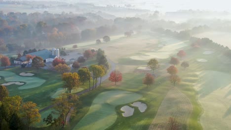 Cinematic-high-aerial-reveals-maintenance-barn-on-golf-course-at-sunrise,-frost-and-fog-in-autumn-sunlight