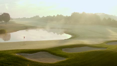 Glide-past-flag-pole,-water-hazard,-sand-traps,-fog-and-frost-in-beautiful-morning-sunrise-light