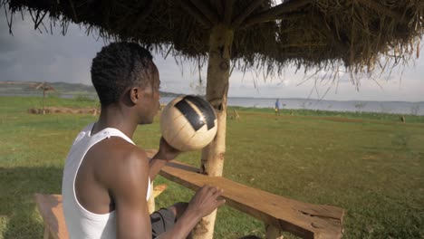 Eine-Nahaufnahme-Eines-Jungen-Afrikanischen-Mannes-Mit-Einem-Fußball,-Der-Sich-Unter-Einer-Strandhütte-Am-Ufer-Des-Viktoriasees-Entspannt