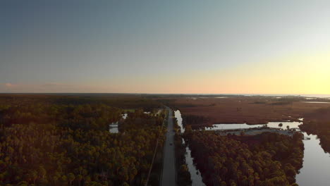 Aerial-view-of-a-long-straight-road-with-a-car-driving-next-to-a-large-forest-and-some-ponds-in-Florida