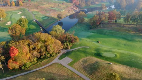 Golf-course-in-autumn,-worker-on-tractor-fertilizers-beautiful-course,-bridge-crossing-river,-orbiting-aerial-shot