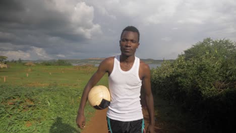 Slow-motion-shot-tracking-from-the-front-of-a-young-African-man-walking-with-a-football-under-his-arm-by-the-stormy-shores-of-Lake-Victoria