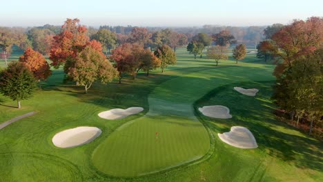 Aerial-turn-at-golf-course-fairway,-flag,-sand-trap-hazards,-autumn-leaves,-sunrise-illuminates-morning-fog