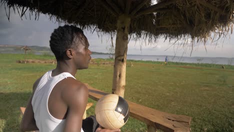 Nahaufnahme-Eines-Jungen-Afrikanischen-Mannes,-Der-Sich-Auf-Einer-Strandhütte-Am-Lake-Victoria-Entspannt,-Während-Er-Mit-Einem-Fußball-Spielt