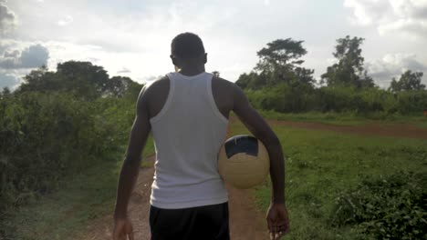 Eine-Kamerafahrt-In-Zeitlupe-Von-Hinten-Eines-Jungen-Afrikanischen-Mannes,-Der-Einen-Fußball-Hält,-Während-Er-Der-Sonne-Entgegengeht
