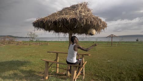 Zeitlupenaufnahme-Hinter-Einem-Afrikanischen-Jugendlichen,-Der-Unter-Einer-Strandhütte-Sitzt,-Während-Er-Mit-Einem-Fußball-Spielt