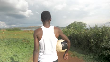 Behind-view-tracking-a-young-African-man-as-he-walks-towards-Lake-Victoria-with-a-football-under-his-arm