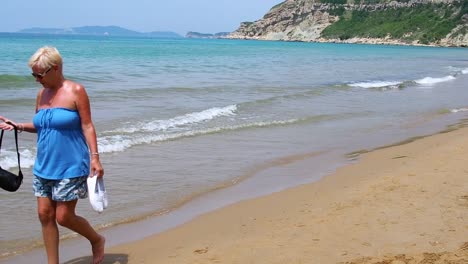 Attractive-middle-aged-female-walking-barefoot-on-beach-feeling-hot-during-midday-sun-Corfu-Greece