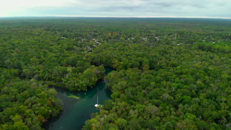 Aerial-footage-of-a-river-cutting-through-a-vast-tropical-forest-with-a-still-sailing-boat-and-some-houses-in-the-distance,-dolly-in-shot
