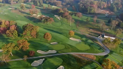 Aerial-in-fall-season,-colorful-leaves-above-golf-course-fairways-during-beautiful-morning-light