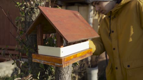 A-Young-Man-Wearing-Yellow-Winter-Jacket,-Checking-On-The-Tiny-Bird-House-On-A-Branch-Of-A-Tree---Close-Up-Shot