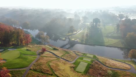 Aerial-pan-of-sunrise-above-golf-course-on-misty-morning,-spectacular-light-falls-on-autumn-tree-leaves,-river,-bridge