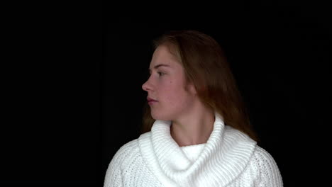 A-close-up-of-a-woman-wearing-a-white-knitted-jumper,-turning-her-head-to-the-side,-on-a-seamless-black-studio-background
