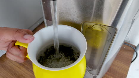 Hand-of-a-girl-adding-hot-water-from-dispenser-in-a-cup-with-dry-green-tea-leaves