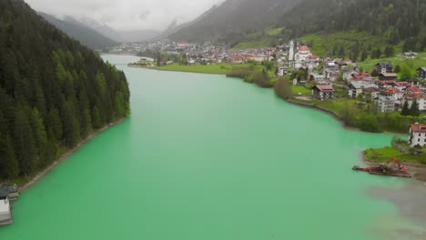 A-green-lake-behind-a-dam-in-the-Italian-Alps,-going-towards-a-town