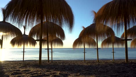 Sonnenaufgang,-Der-Sich-Tief-Auf-Den-Boden-Bewegt,-Schuss-Von-Strohsonnenschirmen-Am-Strand-Von-Marbella,-4K-Gimbal-Kinoansicht