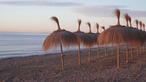 Tiki-Sonnenschirme-Am-Strand-Von-Marbella-Bei-Sonnenaufgang,-Wunderschöne-4k-Spanien-Urlaubsaufnahmen