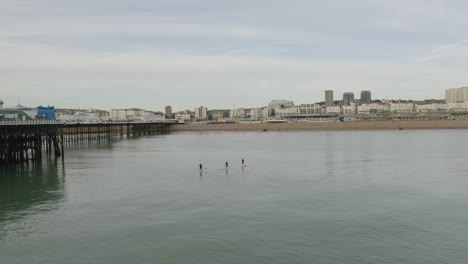 Three-people-on-paddle-boards-heading-for-the-beach-in-Brightonm,-seagulls-flying-past-and-drone-panning-right