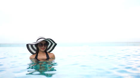 smiling-woman-surrounded-by-crystal-blue-rippling-water-and-bright-sunlight-wearing-a-striped-hat-and-sunglasses