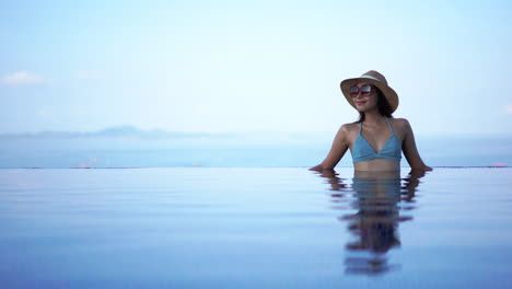 a-lovely-lady-in-a-blue-swimsuit-wearing-a-sun-hat-and-sunglasses-standing-alone-in-a-peaceful-infinity-pool-with-her-reflection-shining-on-calm-clear-blue-water