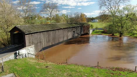 Aerial-push-in-on-covered-bridge-above-muddy-flooded-river-on-beautiful-autumn-day