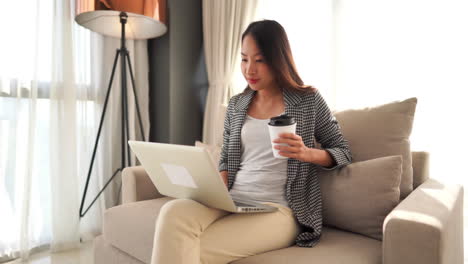 A-pretty-Asian-woman-sitting-on-an-overstuffed-chair-holding-a-coffee-cup-while-typing-on-her-laptop