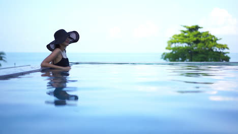 Elegant-lady-wearing-a-black-sun-hat-leaning-peacefully-on-the-edge-of-a-relaxing-infinity-pool-as-crystal-clear-water-ripples-with-her-reflection