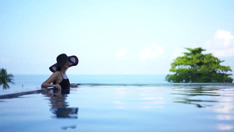 With-the-ocean-horizon-in-the-background,-a-woman-in-a-black-one-piece-bathing-suit-and-black-straw-floppy-sun-hat-lounges-against-the-edge-of-a-resort-pool