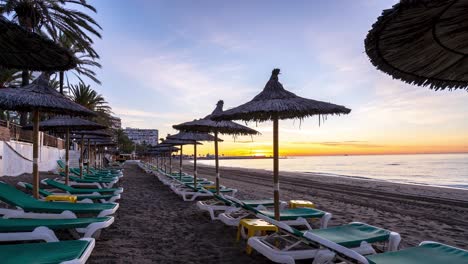 Zeitraffer-Sonnenaufgang-Am-Strand-Von-Marbella-Mit-Sonnenliegen-Und-Sonnenschirmen,-Urlaubsziel-4k