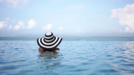 A-woman-with-her-back-to-the-camera-wearing-a-floppy-black-and-white-sunhat-is-neck-deep-in-a-resort-swimming-pool-looking-out-to-the-ocean